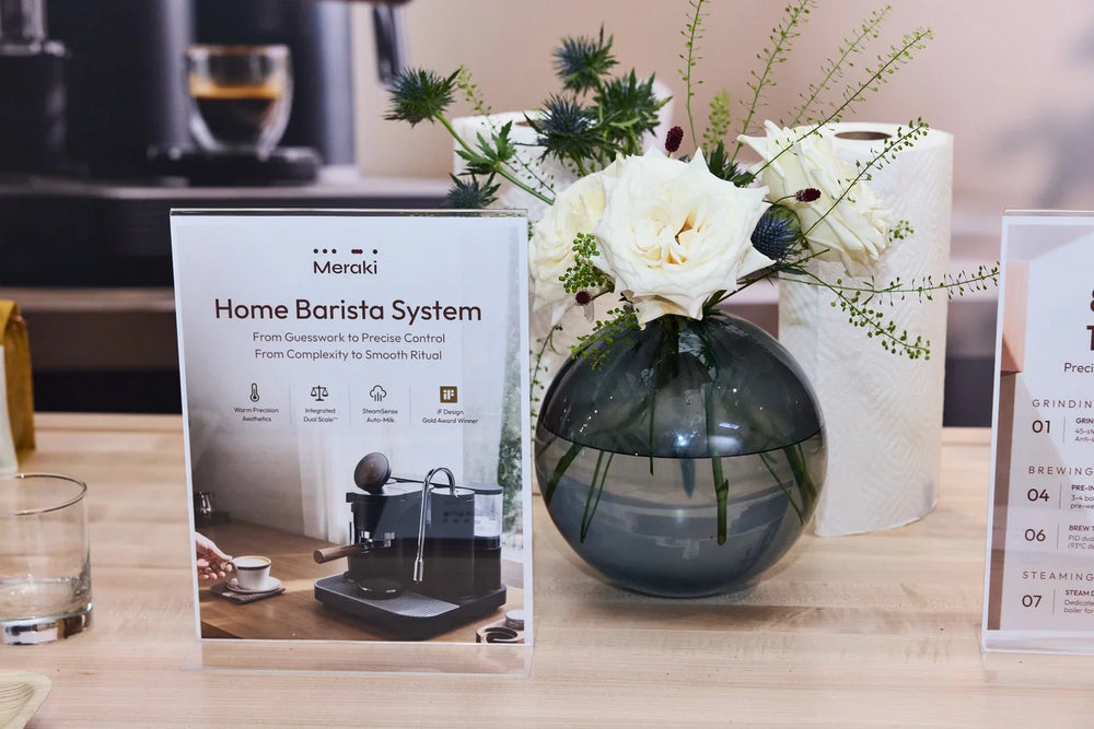 Meraki „Home Barista System“-Produktaufsteller neben weißen Blumen in einer Glasvase auf der Holztheke beim New York Experience Event.