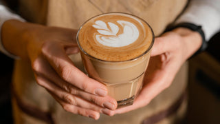 Hände halten ein frisch zubereitetes Latte-Art-Kaffeegetränk im Glas