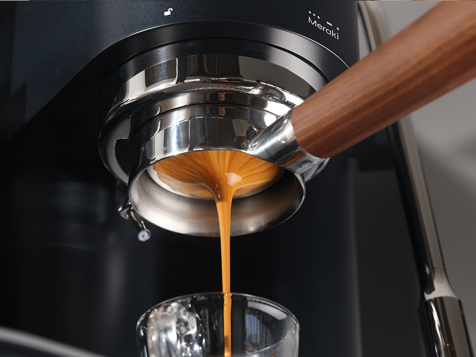 Espresso fließt aus einem bodenlosen Siebträger mit Holzgriff in ein Glas an einer schwarzen Meraki-Espressomaschine.