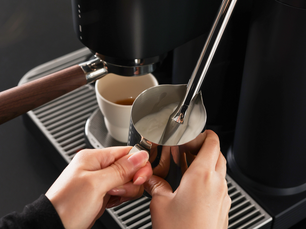 Hände halten ein Milchkännchen unter die Dampflanze; im Hintergrund steht eine Espressotasse auf der Abtropfschale.