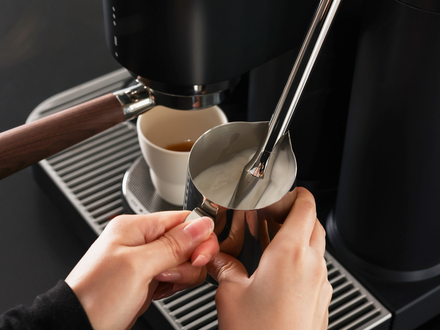 Hände halten ein Milchkännchen unter die Dampflanze; im Hintergrund steht eine Espressotasse auf der Abtropfschale.